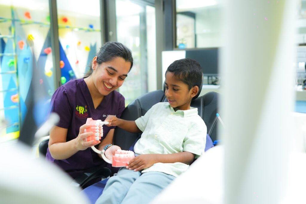 A smiling dental professional shows a young boy how to brush teeth using a large tooth model and toothbrush in a brightly lit dental clinic.