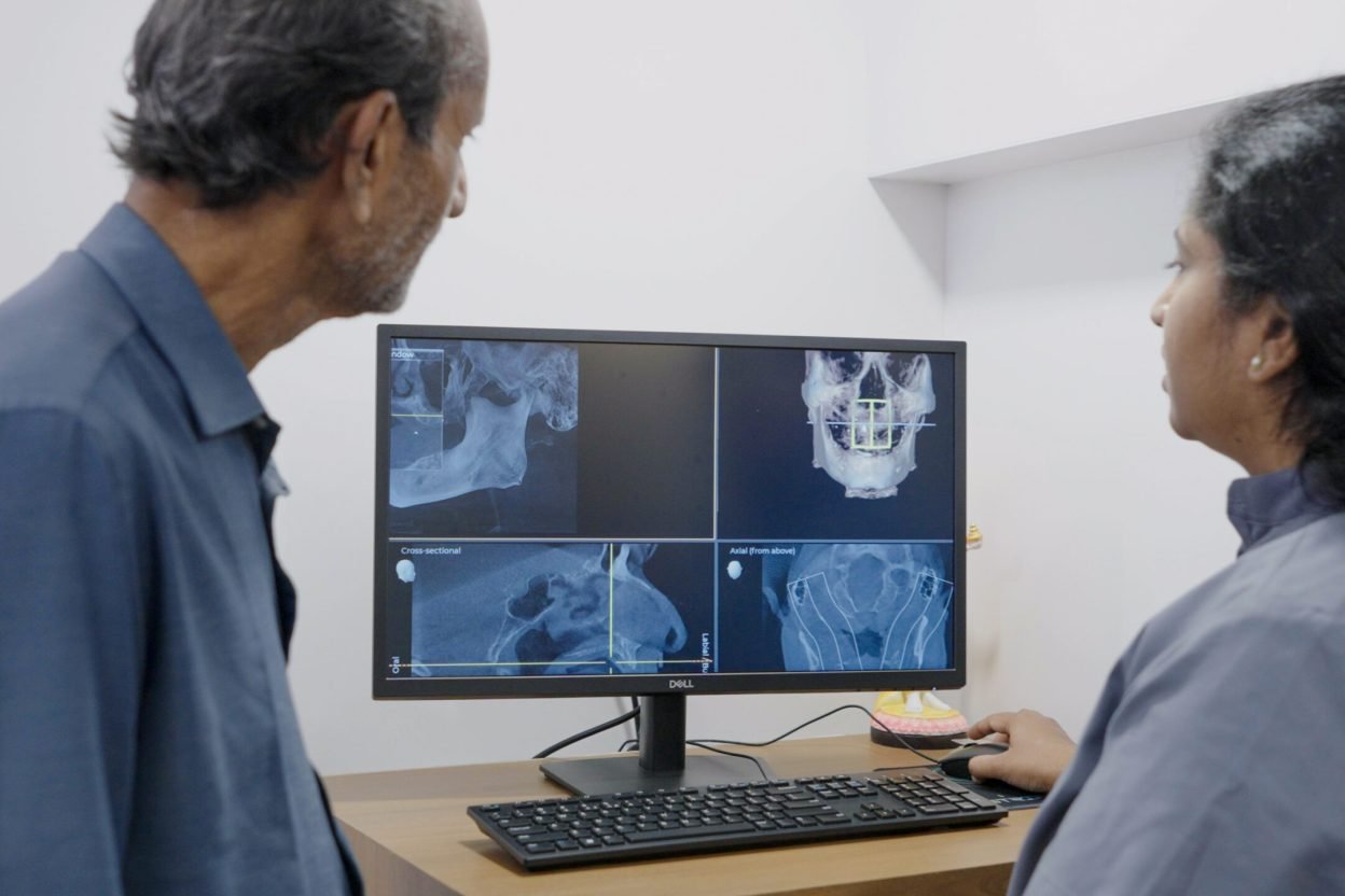 A man and a woman look at dental X-ray images displayed on a computer monitor in a medical office. The woman points to the screen, possibly explaining the images to the man.